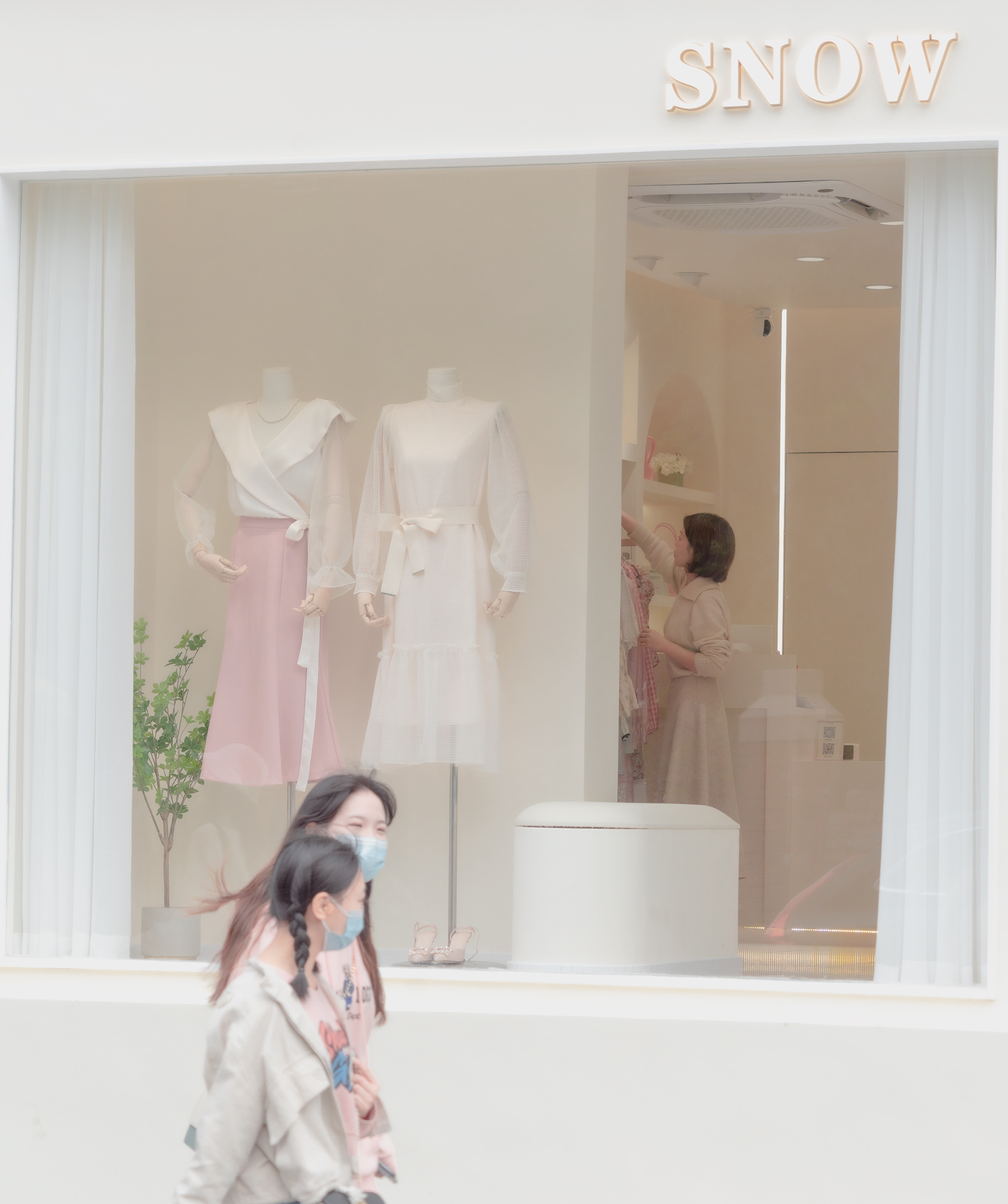two girls passing by a consice clothes shop,the proprietress is tidying clothes through the window